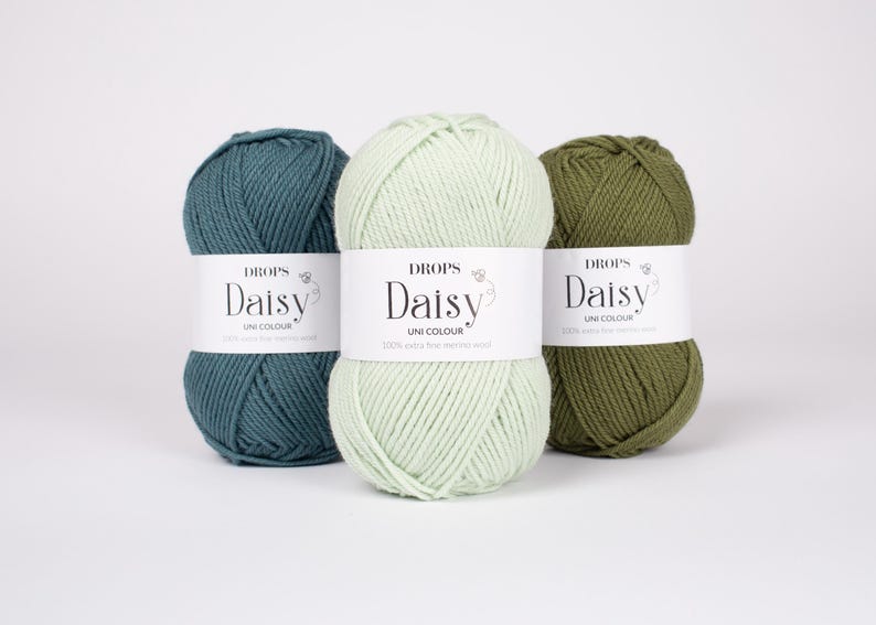 May include: Three skeins of Drops Daisy yarn in teal, light mint green, and olive green. Each skein features a white paper label with the text "DROPS Daisy UNI COLOUR 100% extra fine merino wool" printed in black, centered on the yarn.