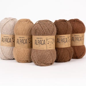 May include: Five skeins of Drops Alpaca yarn arranged in a row, featuring a gradient of neutral earth tones from light beige to dark brown. Each ball of yarn is wrapped in a tan paper label with the text Drops Alpaca and a small alpaca illustration.