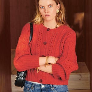 May include: A woman wearing a red cardigan sweater with a round neckline and button closure. The sweater has a cable knit design on the sleeves and a ribbed hem. She is wearing blue jeans, gold jewelry, and carrying a black purse.