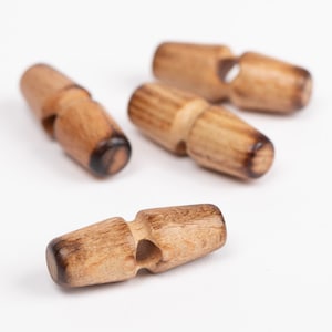 May include: A collection of four wooden toggle buttons. Each button is a light brown color with a darker, burnt-looking end. The buttons have a cylindrical shape with a central hole for sewing. The buttons are scattered on a white surface.