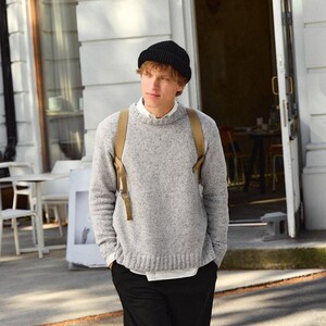 May include: A man wearing a grey sweater, black trousers, and a black beanie. He is standing outside a building with a door behind him.