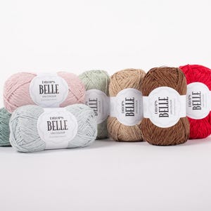 May include: A collection of colorful yarn skeins, including shades of pink, green, tan, brown, and red. Each skein has a white label with the text "BELLE" and "UNI COLOUR". The yarn is arranged on a white surface.