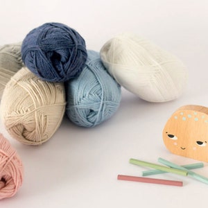 Puede incluir: Seis ovillos de lana en varios colores, incluyendo azul claro, beige, rosa claro, blanco y azul oscuro, est&aacute;n dispuestos sobre una superficie blanca. Una ballena de juguete de madera con una cara pintada y algunos palitos de madera tambi&eacute;n est&aacute;n sobre la superficie.