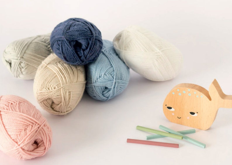 Puede incluir: Seis ovillos de lana en varios colores, incluyendo azul claro, beige, rosa claro, blanco y azul oscuro, est&aacute;n dispuestos sobre una superficie blanca. Una ballena de juguete de madera con una cara pintada y algunos palitos de madera tambi&eacute;n est&aacute;n sobre la superficie.