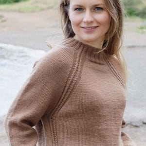May include: A light brown long-sleeved knitted sweater with a ribbed crew neckline and textured raglan seams. The garment features a solid color knit pattern throughout. The image includes the text DROPS DESIGN and www.garnstudio.com.