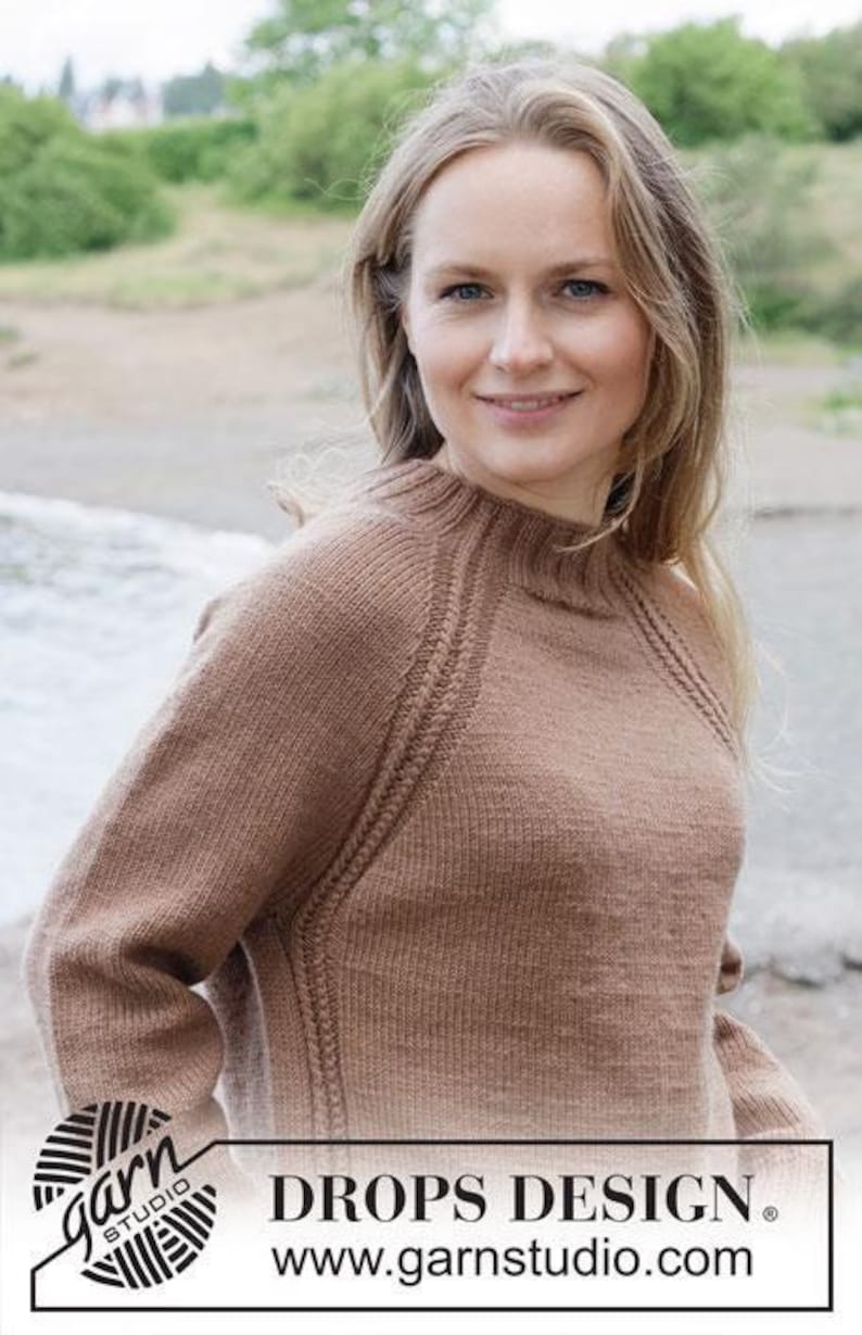 May include: A light brown long-sleeved knitted sweater with a ribbed crew neckline and textured raglan seams. The garment features a solid color knit pattern throughout. The image includes the text DROPS DESIGN and www.garnstudio.com.