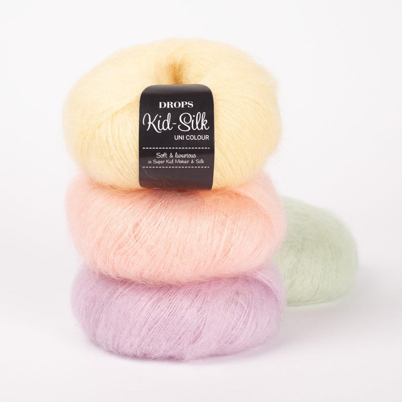 May include: A stack of four balls of Drops Kid-Silk yarn in pastel colors: yellow, peach, lavender, and light green. The top ball has a black label with the brand name and product details. The yarn appears soft and luxurious.