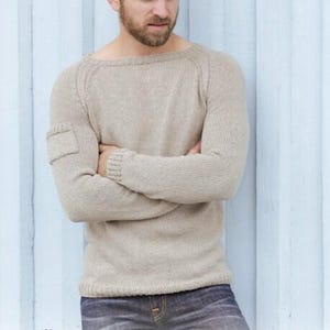 May include: A light beige sweater with a crew neck and a small pocket on the left sleeve. The sweater is paired with blue jeans. The image includes the text "DROPS DESIGN" and "www.garnstudio.com".