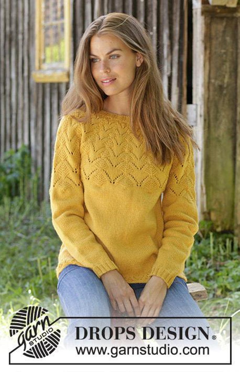 Puede incluir: Un su&eacute;ter de color amarillo mostaza de manga larga con cuello redondo y un patr&oacute;n de encaje de chevr&oacute;n decorativo en la parte superior del pecho y los hombros. La prenda se combina con jeans azules. La imagen incluye el texto DROPS DESIGN.