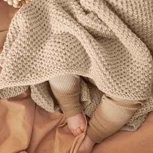 May include: A beige knitted baby blanket with a textured pattern. The blanket is draped over a baby's legs, with the baby's feet visible.