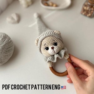 Op de afbeelding: Gehaakte amigurumi berenrammelaar met een houten ring. De beer draagt ​​een grijze gehaakte muts en heeft een gerimpelde kraag. Het speelgoed is gemaakt van beige en grijs garen.