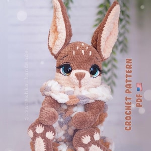 Puede incluir: Un conejo de ganchillo hecho a mano en tonos marrones, beige y blanco. El conejo tiene ojos grandes y expresivos con largas pestañas y un cuerpo esponjoso y texturizado. El texto "CROCHET PATTERN PDF" es visible en el lado derecho de la imagen.