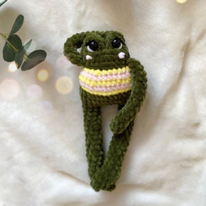 May include: A handmade green crochet frog doll with large black eyes and a yellow and pink striped belly. The frog has long legs and arms, and is posed with one arm raised. The doll is made of soft, plush yarn.