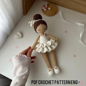 Könnte beinhalten: Häkelanleitung für eine Ballerina-Puppe mit braunen Haaren, einem weißen Tutu und weißen Ballettschuhen. Die Anleitung ist in englischer Sprache verfügbar und beinhaltet einen PDF-Download.