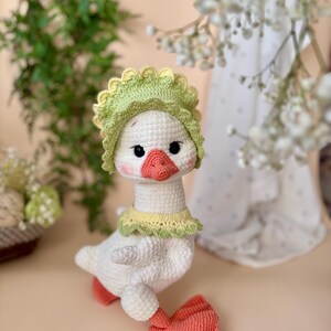 May include: A handmade crochet goose doll with a white body, wearing a green and yellow bonnet and collar. The goose has a peach-colored beak and feet, and black eyes. The doll is a charming, whimsical creation.