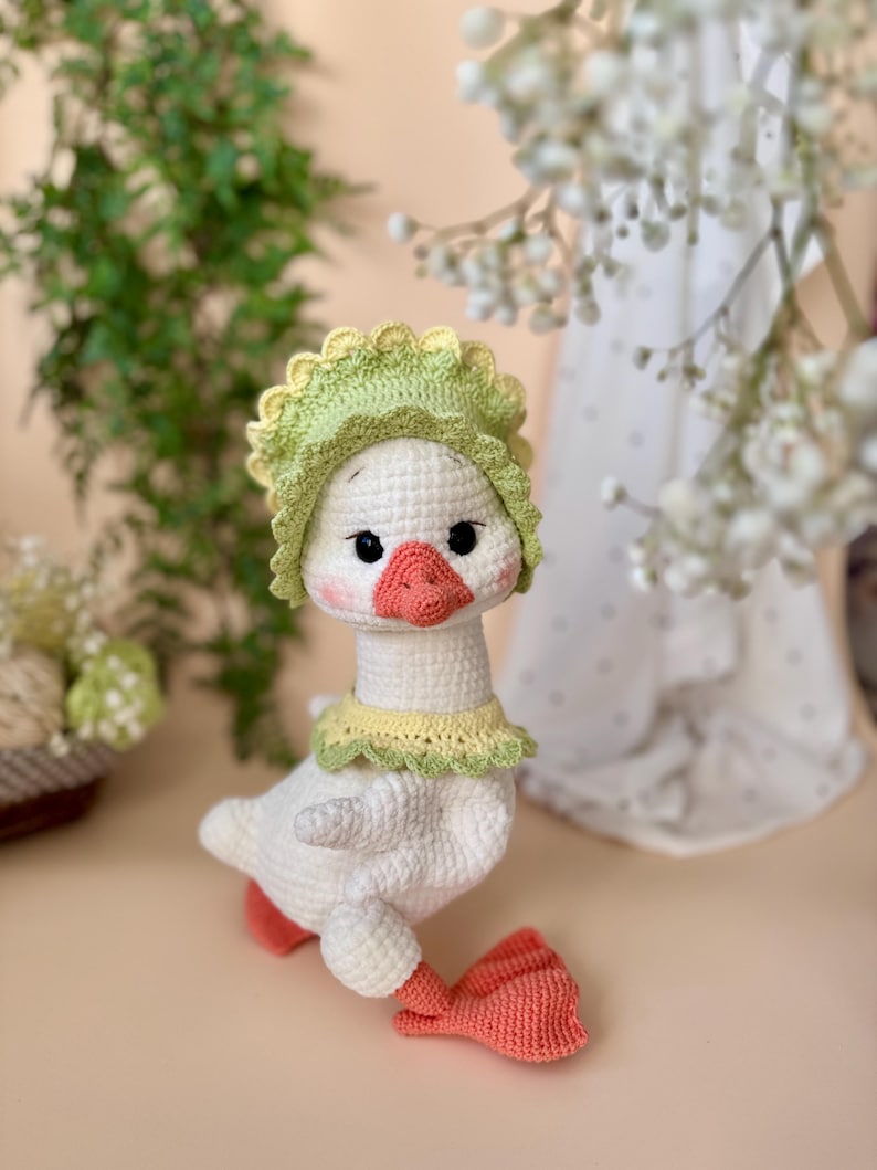 May include: A handmade crochet goose doll with a white body, wearing a green and yellow bonnet and collar. The goose has a peach-colored beak and feet, and black eyes. The doll is a charming, whimsical creation.
