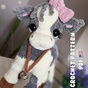 Può includere: Un peluche a forma di mucca realizzato in filato grigio e crema con un fiocco rosa sulla testa, ciglia lunghe e una collana di filato marrone con una piccola campanella argentata. Il testo CROCHET PATTERN PDF è visibile a destra con una bandiera.