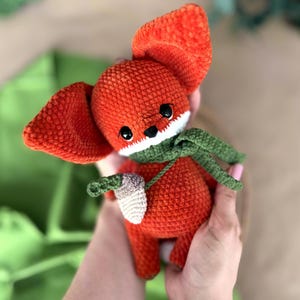 May include: A handmade orange crochet fox doll with large ears, black eyes, and a white muzzle. The fox wears a green scarf and holds a small, off-white and brown acorn. A charming, soft toy.