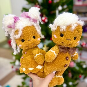 Puede incluir: Dos mu&ntilde;ecas de jengibre de crochet, una con un lazo rosa y la otra con un lazo marr&oacute;n, se toman de la mano. Ambas llevan gorros de piel blanca y tienen ojos marrones. Las mu&ntilde;ecas est&aacute;n delante de un &aacute;rbol de Navidad.