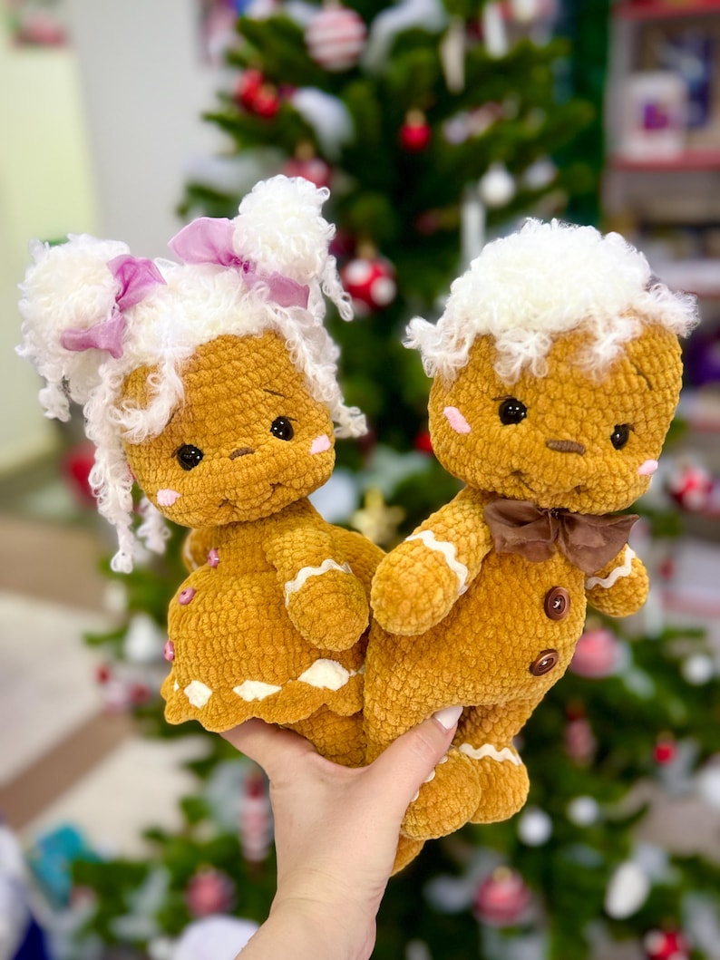 Puede incluir: Dos mu&ntilde;ecas de jengibre de crochet, una con un lazo rosa y la otra con un lazo marr&oacute;n, se toman de la mano. Ambas llevan gorros de piel blanca y tienen ojos marrones. Las mu&ntilde;ecas est&aacute;n delante de un &aacute;rbol de Navidad.
