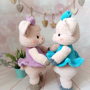Op de afbeelding: Twee gehaakte varkens knuffels die jurken dragen. Het varken links draagt een lavendelkleurige jurk en het varken rechts draagt een turquoise jurk. Beide varkens hebben grijze gehaakte schoenen.