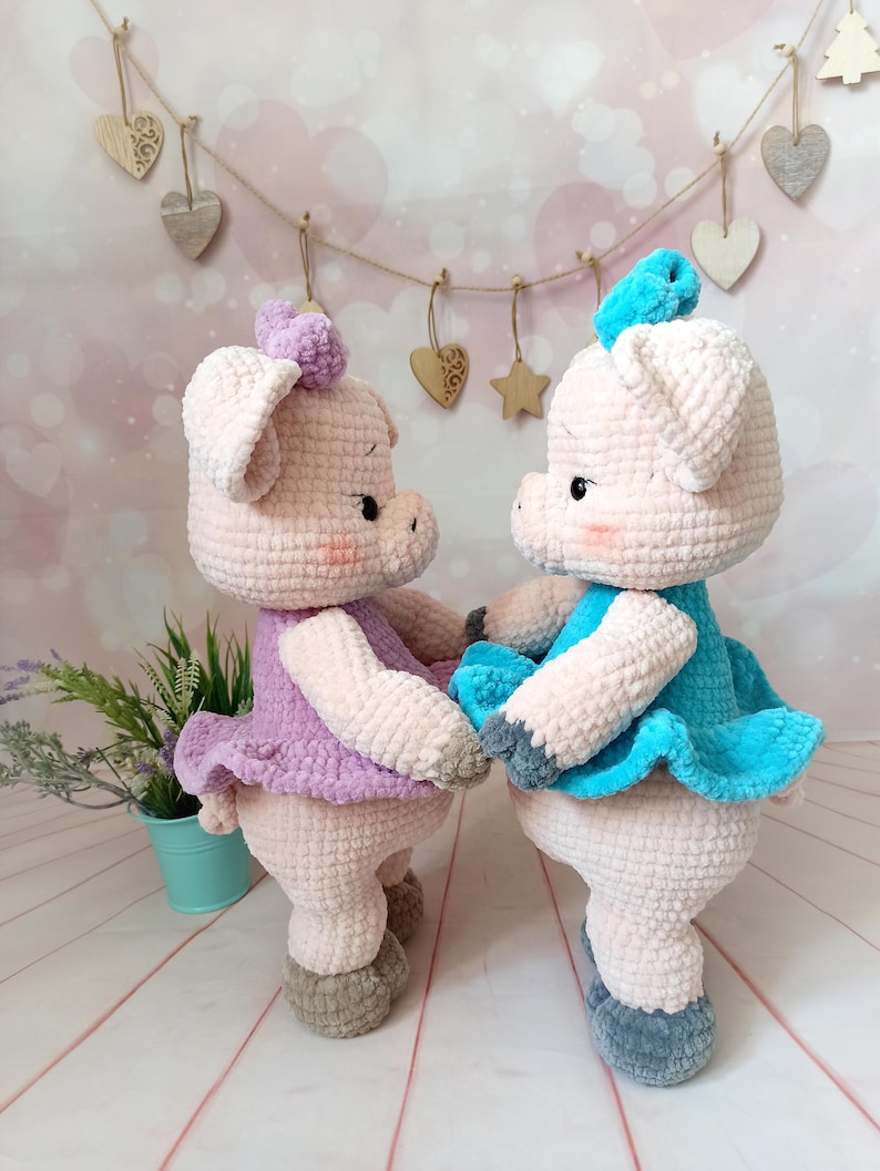 Op de afbeelding: Twee gehaakte varkens knuffels die jurken dragen. Het varken links draagt een lavendelkleurige jurk en het varken rechts draagt een turquoise jurk. Beide varkens hebben grijze gehaakte schoenen.