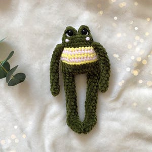 May include: A handmade green crochet frog plush toy with a yellow and pink striped belly. The frog has large black eyes and long, dangling legs. The plush is made of soft, textured yarn.