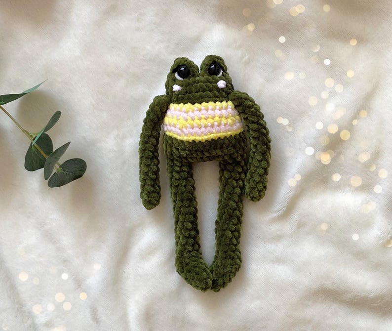 May include: A handmade green crochet frog plush toy with a yellow and pink striped belly. The frog has large black eyes and long, dangling legs. The plush is made of soft, textured yarn.