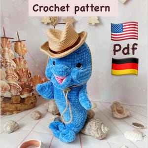 May include: A blue crocheted dolphin wearing a tan cowboy hat and a rope necklace. The dolphin has a pink mouth and black eyes. The image includes the text "Crochet pattern" and a PDF icon with the American and German flags.