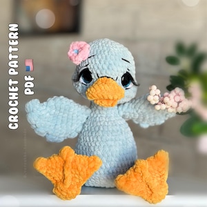 May include: A light blue crocheted duck plush toy with a bright orange beak and webbed feet. The duck features large dark eyes with long eyelashes, a small pink flower accessory on its head, and holds a cluster of tiny pink berries.