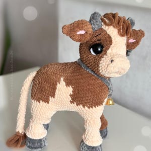 May include: A plush cow toy made of brown and cream yarn with a soft, textured finish. The cow features large black eyes with long eyelashes, small grey horns, and a grey collar around its neck with a small, dangling gold-colored metal bell.