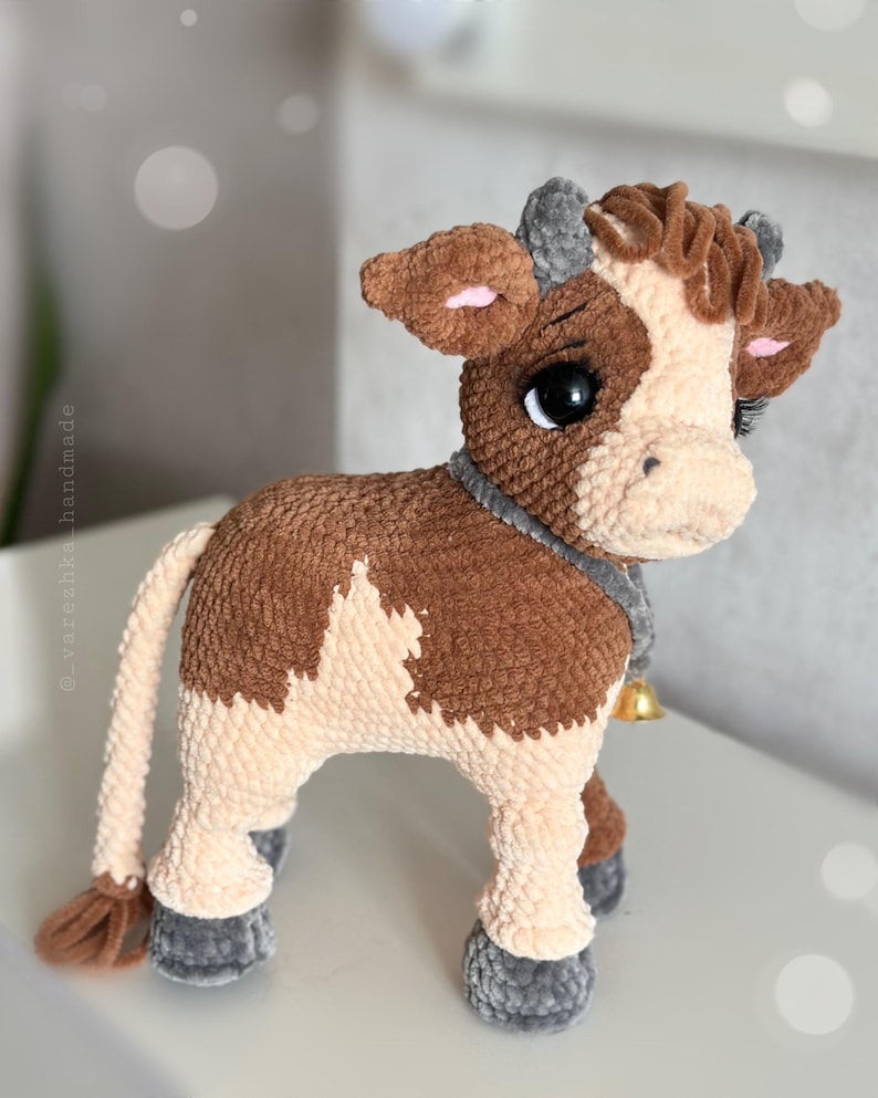 May include: A plush cow toy made of brown and cream yarn with a soft, textured finish. The cow features large black eyes with long eyelashes, small grey horns, and a grey collar around its neck with a small, dangling gold-colored metal bell.
