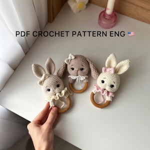 May include: Three handmade crochet bunny rattles in beige, tan, and cream. Each bunny has a wooden ring and a decorative bow or ruffle. The text "PDF CROCHET PATTERN ENG" is visible.
