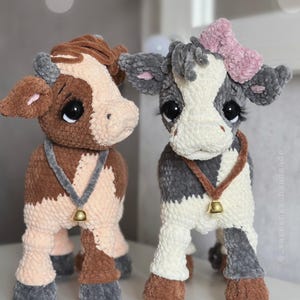 May include: Two plush cow toys made of soft yarn. The left cow is brown and cream with a gray collar and bell. The right cow is gray and white with a pink bow, a brown collar with a bell, and long eyelashes. The text _varezhka_handmade is visible.