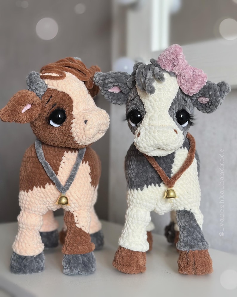 May include: Two plush cow toys made of soft yarn. The left cow is brown and cream with a gray collar and bell. The right cow is gray and white with a pink bow, a brown collar with a bell, and long eyelashes. The text _varezhka_handmade is visible.