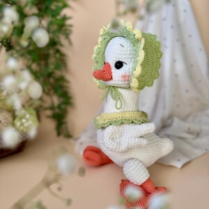 May include: A handmade white crochet goose wearing a green and yellow bonnet and collar. The goose has an orange beak and feet, and black eyes. The plush toy is a whimsical, soft sculpture.