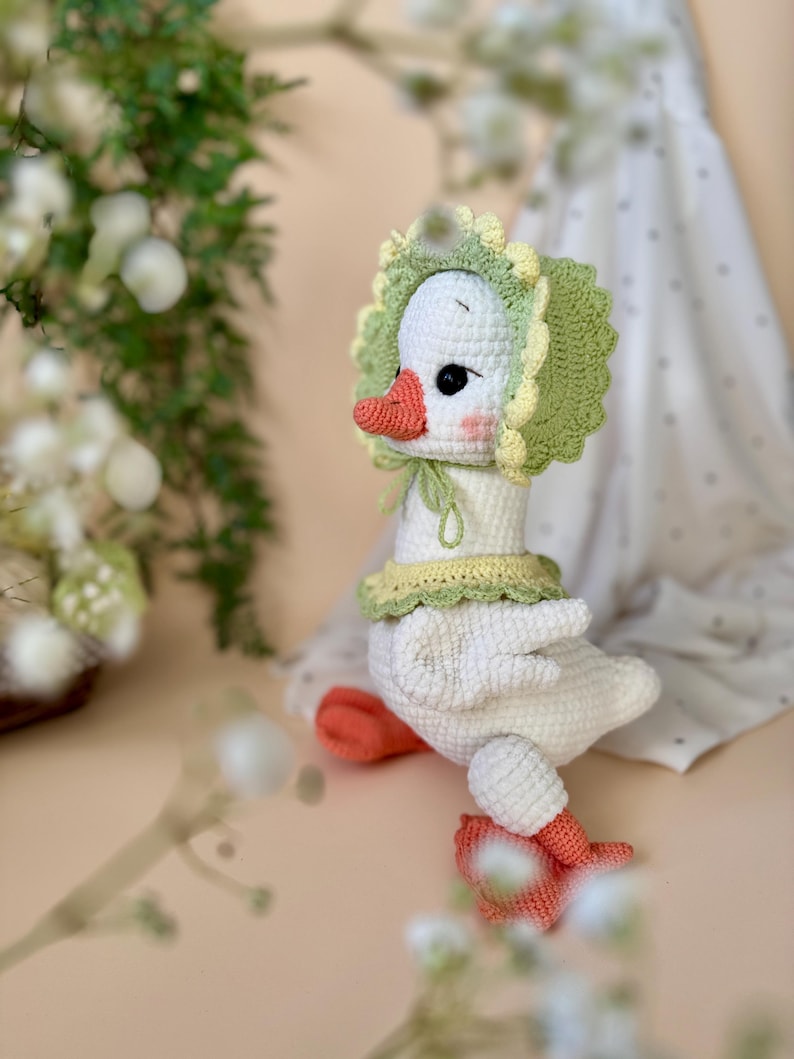May include: A handmade white crochet goose wearing a green and yellow bonnet and collar. The goose has an orange beak and feet, and black eyes. The plush toy is a whimsical, soft sculpture.