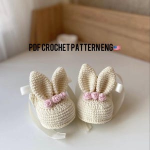 Könnte beinhalten: Ein Paar gehäkelte Hasenschuhe mit rosa Blumenakzenten. PDF CROCHET PATTERN ENG ist auf dem Bild geschrieben.