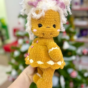 Puede incluir: Una mu&ntilde;eca de jengibre de crochet con cabello blanco rizado, lazos rosas y un vestido blanco y marr&oacute;n. La mu&ntilde;eca est&aacute; de pie frente a un &aacute;rbol de Navidad verde.