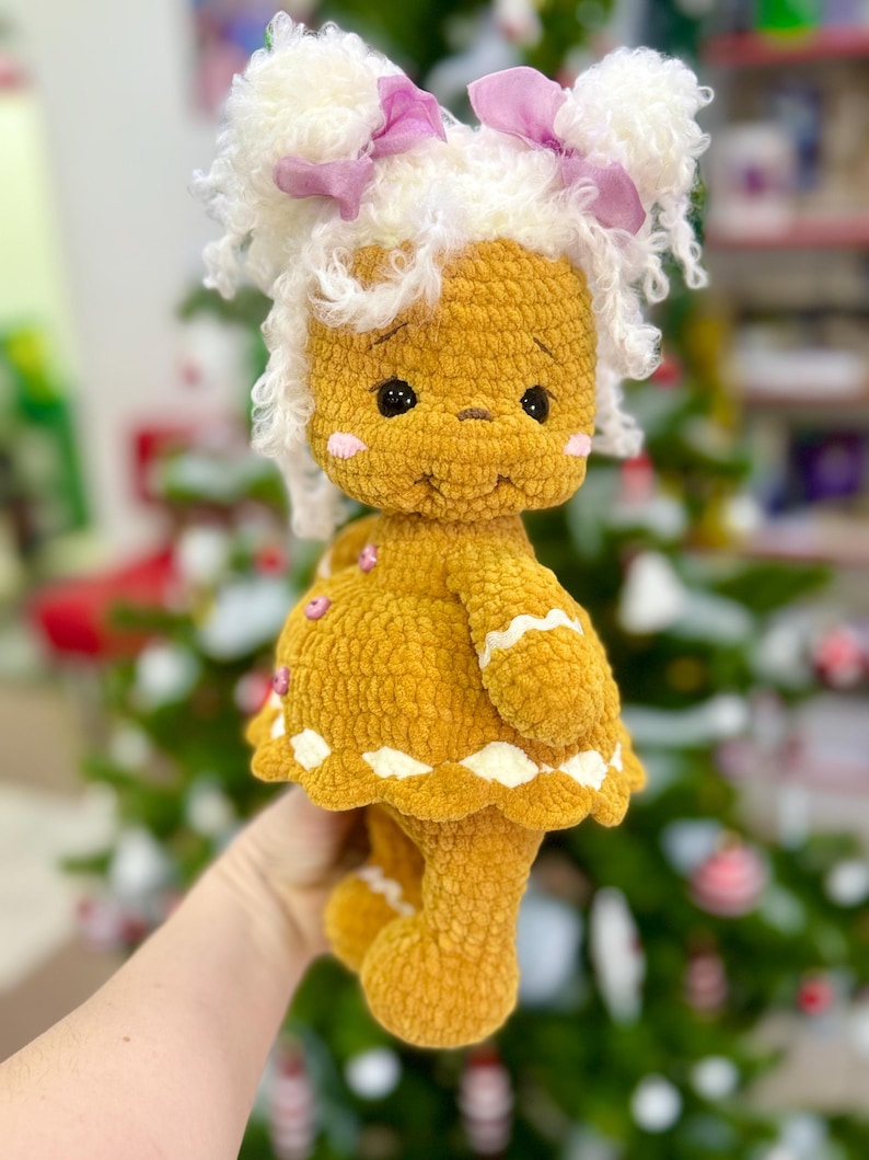 Puede incluir: Una mu&ntilde;eca de jengibre de crochet con cabello blanco rizado, lazos rosas y un vestido blanco y marr&oacute;n. La mu&ntilde;eca est&aacute; de pie frente a un &aacute;rbol de Navidad verde.