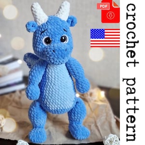 Op de afbeelding: Een blauw gehaakt draken amigurumi speelgoed met witte hoorns en ogen. Het speelgoed staat op een lichtbruin oppervlak. De afbeelding bevat de tekst "crochet pattern" en een downloadpictogram.