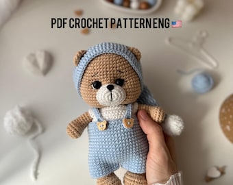 May include: A crocheted teddy bear wearing a blue and white outfit with a crocheted hat. The bear has brown eyes and a brown nose. The bear is holding a white crocheted blanket in its right paw.