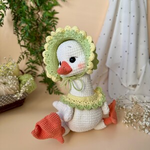 May include: A handmade white crochet goose wearing a green and yellow bonnet and collar. The goose has a peach-colored beak and feet, with rosy cheeks. The goose is sitting down, and the crochet work is detailed.