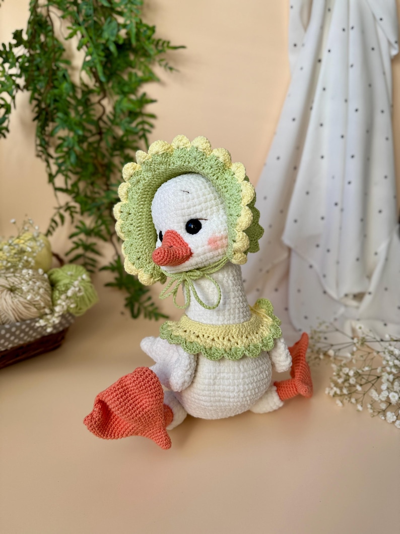 May include: A handmade white crochet goose wearing a green and yellow bonnet and collar. The goose has a peach-colored beak and feet, with rosy cheeks. The goose is sitting down, and the crochet work is detailed.