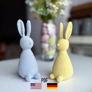 May include: Two crocheted bunny figurines, one light blue and one yellow, sit side-by-side on a white surface. The bunnies have long ears and are in a sitting position. The image includes a vase of flowers in the background.