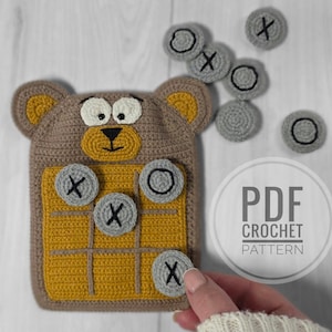 Könnte beinhalten: Gehäkeltes Tic-Tac-Toe-Spiel in Form eines Bären. Das Spielbrett ist braun und gelb, mit grauen X- und O-Spielsteinen. Das Bild enthält den Text "PDF CROCHET PATTERN".