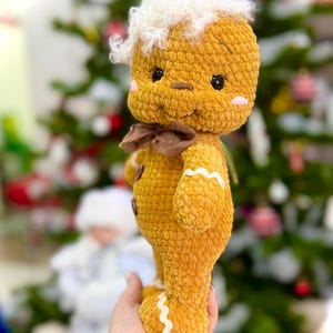 Puede incluir: Un juguete de jengibre de crochet con pelo blanco y esponjoso, un lazo marr&oacute;n y detalles de glaseado blanco. El juguete se sostiene en una mano contra un fondo borroso de un &aacute;rbol de Navidad.