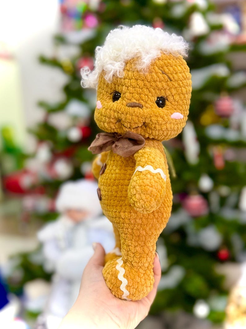 Puede incluir: Un juguete de jengibre de crochet con pelo blanco y esponjoso, un lazo marr&oacute;n y detalles de glaseado blanco. El juguete se sostiene en una mano contra un fondo borroso de un &aacute;rbol de Navidad.