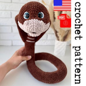 May include: A crocheted brown cobra with a light beige face and blue eyes. The snake has white stripes on its neck. The image includes a PDF icon and the text "Crochet pattern".