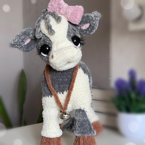 May include: A plush cow toy made of soft gray and white yarn with a pink bow on its head. The cow has large black eyes with long eyelashes, a brown collar with a silver bell, and brown hooves. The text @vareshka_handmade is visible on the side.